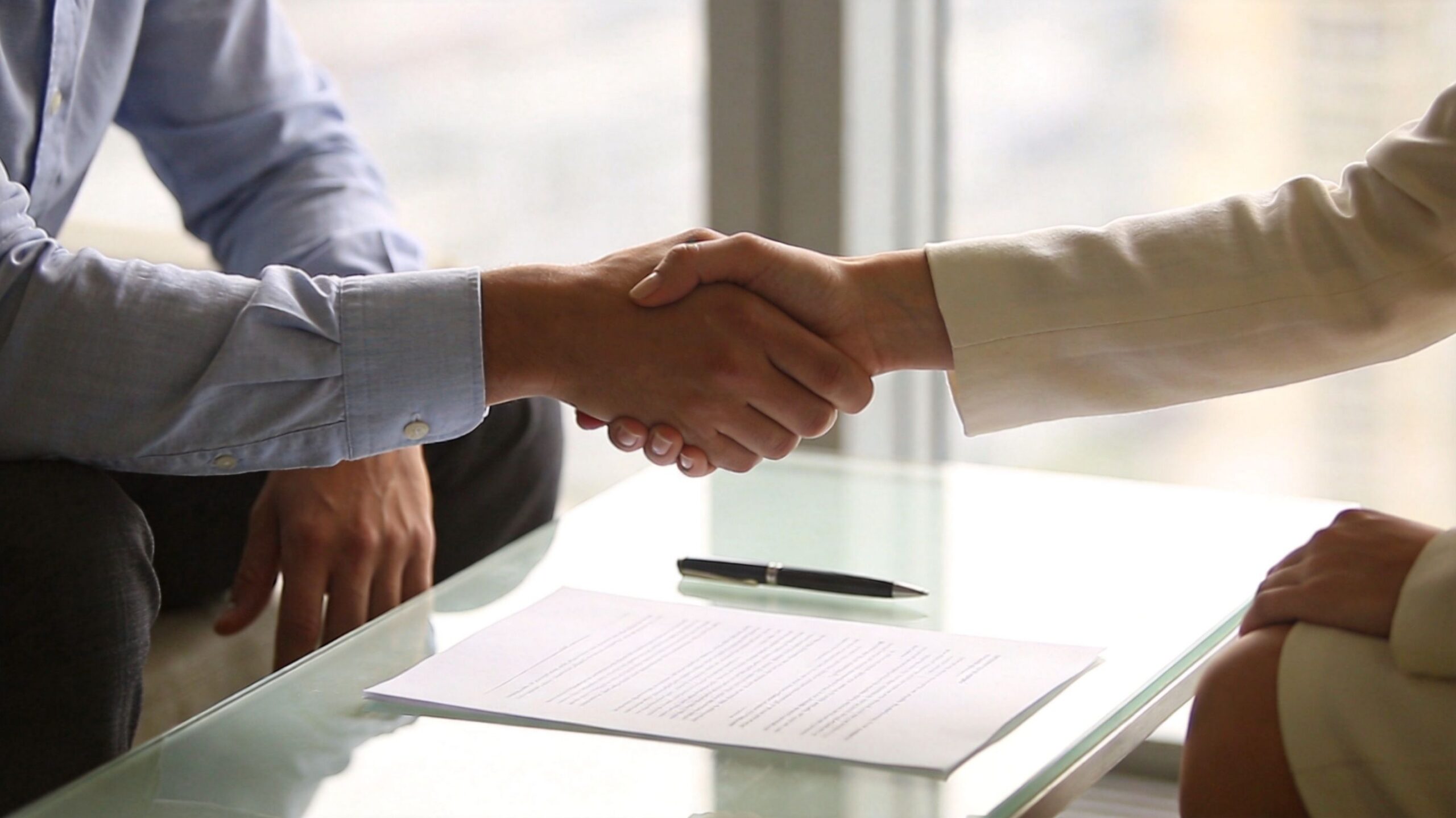 A professional man and woman shaking hands over a business contract on a glass desk.